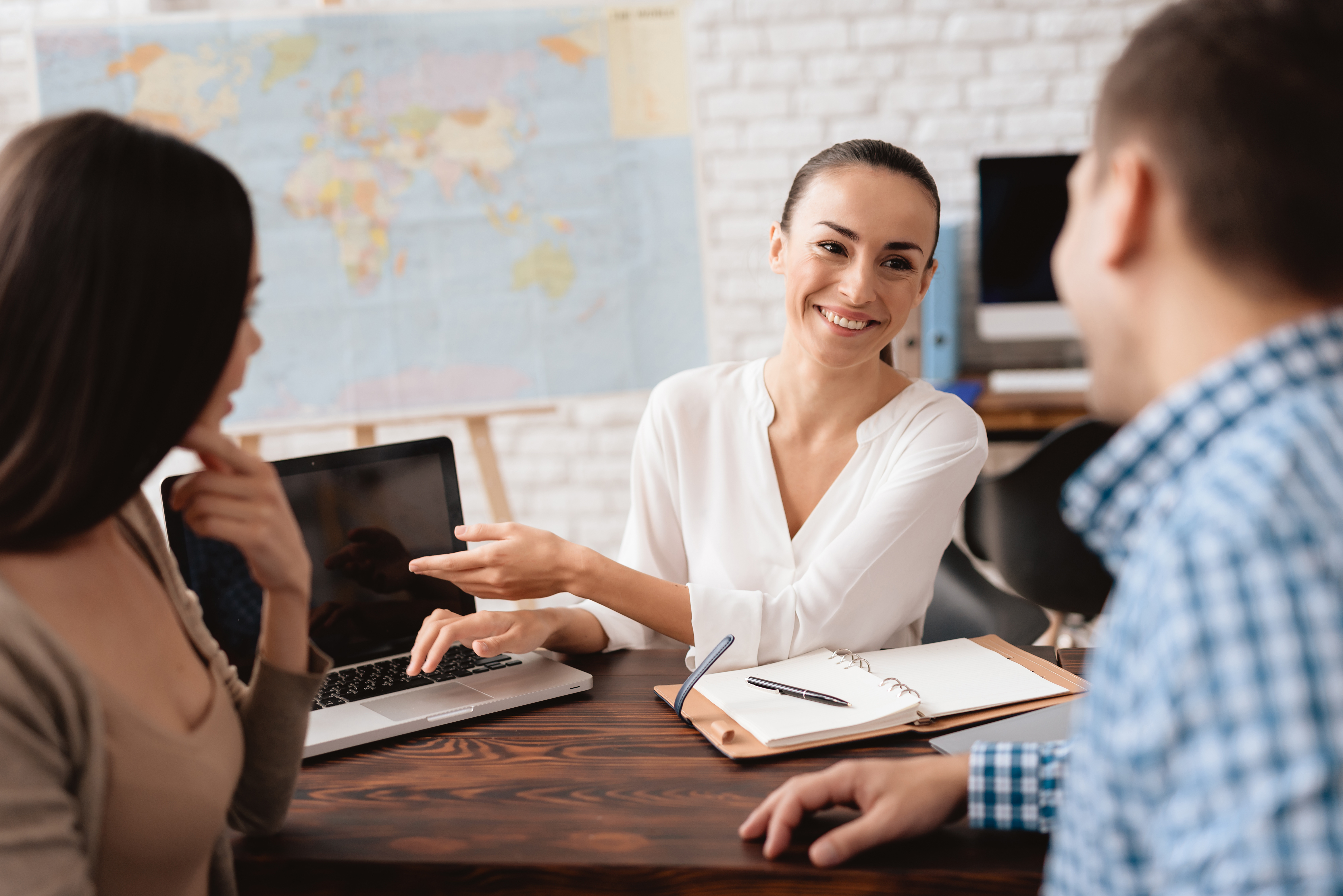 A young man and a woman came to the travel agency. They want to go on a trip during their holidays. The girl agent offers them different countries. She shows it on the laptop.